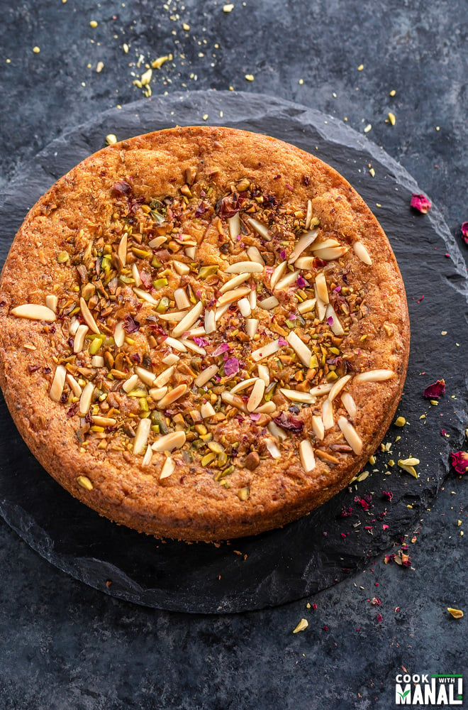 a round eggless mawa cake decorated with nuts and rose petals on top with few spoons placed on the side