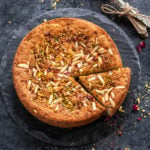 a round eggless mawa cake decorated with nuts and rose petals on top with few spoons placed on the side
