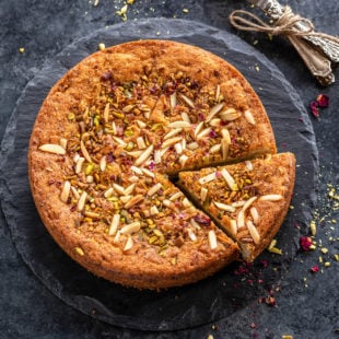 a round eggless mawa cake decorated with nuts and rose petals on top with few spoons placed on the side