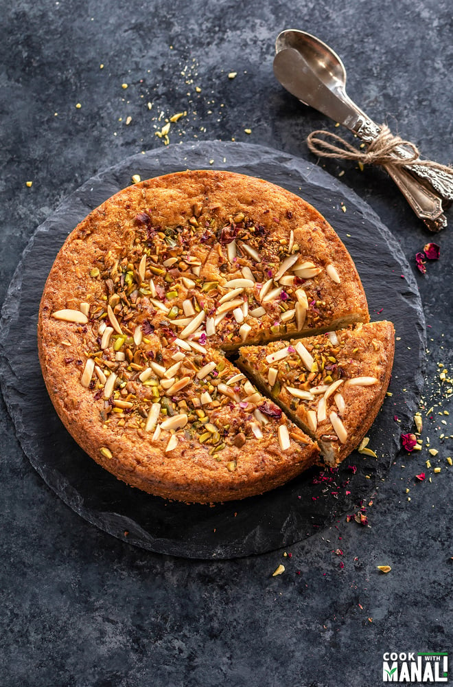 a round eggless mawa cake decorated with nuts and rose petals on top with few spoons placed on the side