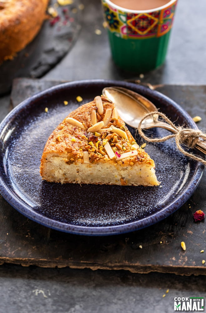 slice of a cake placed in a blue plate with few spoons on side and a glass of chai in the background