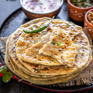 stack of paneer paratha with green chili on the side and bowls of yogurt, pickle in the background