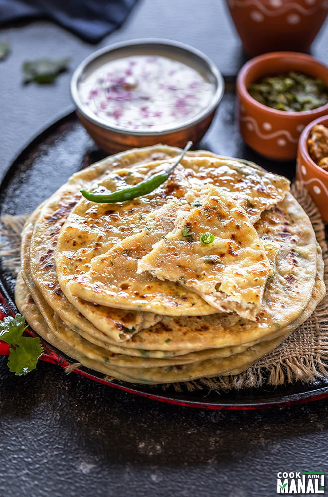 stack of paneer paratha with green chili on the side and bowls of yogurt, pickle in the background