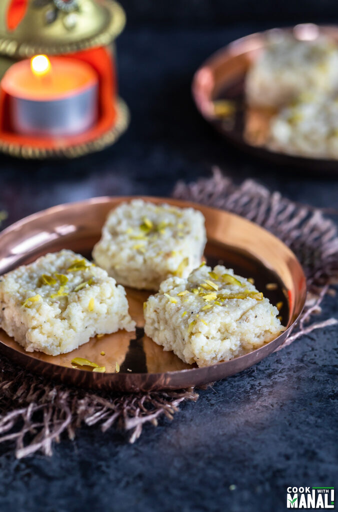 3 pieces of kalakand served in a copper plate with a diya placed in the background