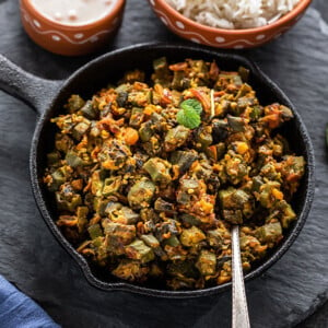 Bhindi Masala in a small iron skillet with a spoon with bowl of rice in the background