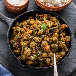 Bhindi Masala in a small iron skillet with a spoon with bowl of rice in the background
