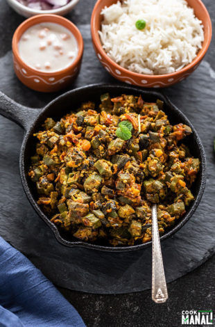 Bhindi Masala in a small iron skillet with a spoon with bowl of rice in the background