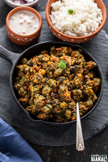 Bhindi Masala in a small iron skillet with a spoon with bowl of rice in the background