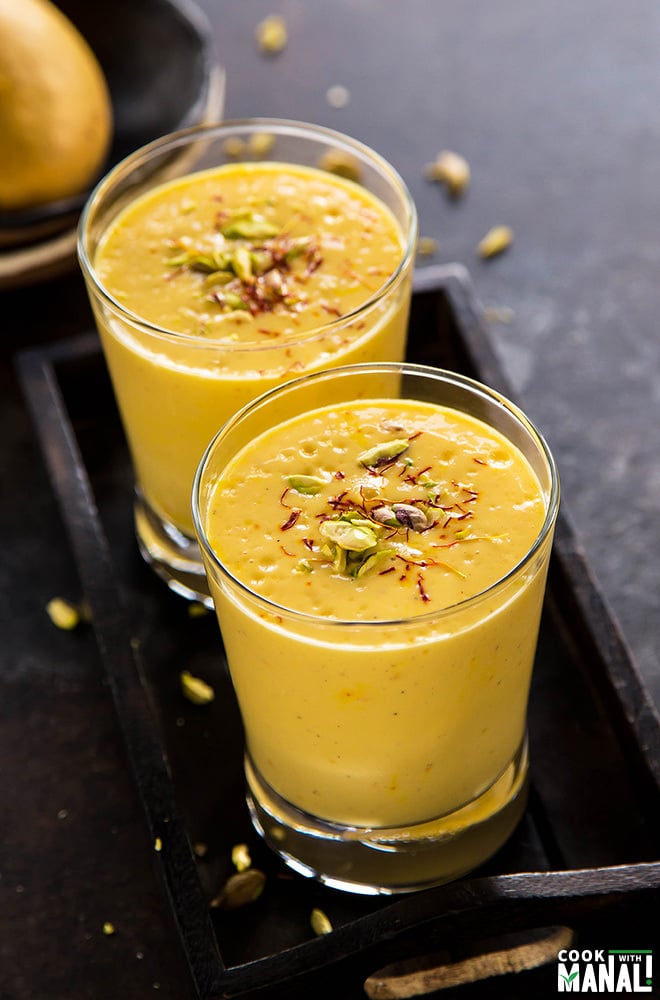 2 glasses of mango lassi garnished with pistachios and a fresh mango in the background