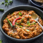 baingan bharta served in a black bowl garnished with green chilies and a spoon on the side