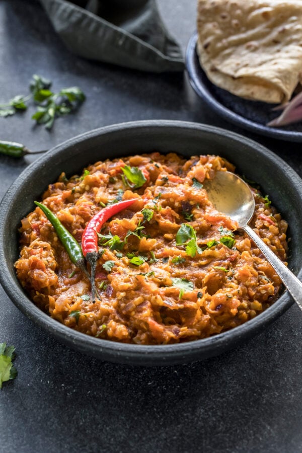 baingan bharta served in a black bowl garnished with green chilies and a spoon on the side