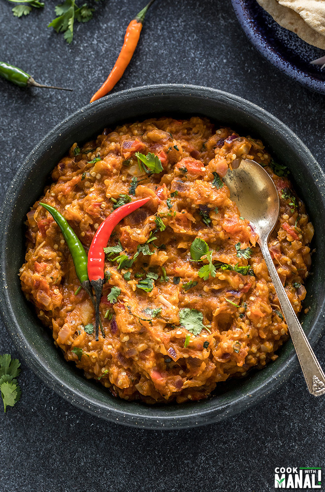 baingan bharta served in a black bowl garnished with green chilies and a spoon on the side