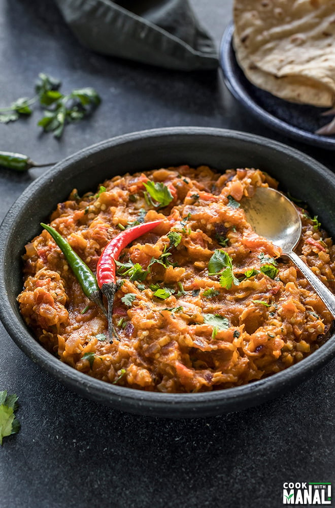 baingan bharta served in a black bowl garnished with green chilies and a spoon on the side