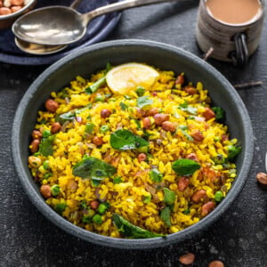 poha served in a black bowl garnished with cilantro and lemon wedges with cups of chai placed in the background