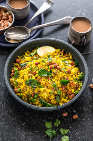 poha served in a black bowl garnished with cilantro and lemon wedges with cups of chai placed in the background