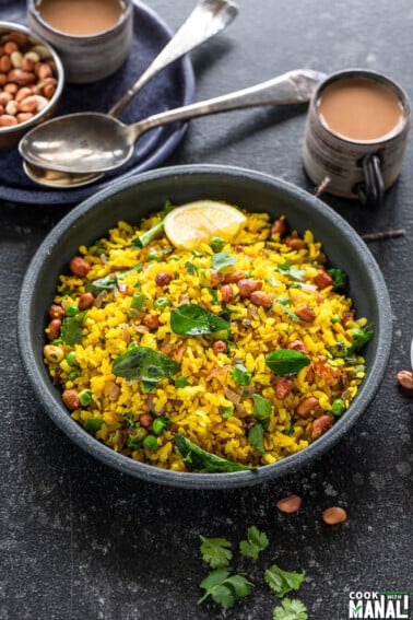poha served in a black bowl garnished with cilantro and lemon wedges with cups of chai placed in the background