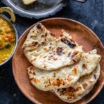 3 pieces of homemade naan placed one over another on a brown plate with a curry placed on the side and a slab of butter in the background