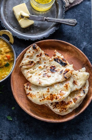 3 pieces of homemade naan placed one over another on a brown plate with a curry placed on the side and a slab of butter in the background