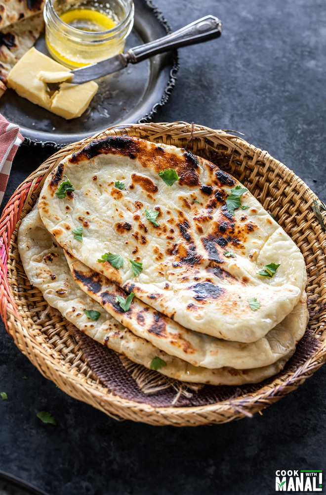 homemade naan stacked and placed on a round bamboo bowl with a slab of butter in the background