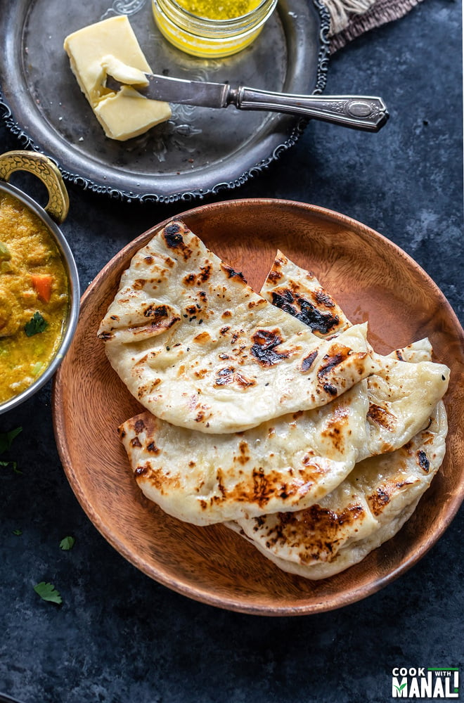3 pieces of homemade naan placed one over another on a brown plate with a curry placed on the side and a slab of butter in the background