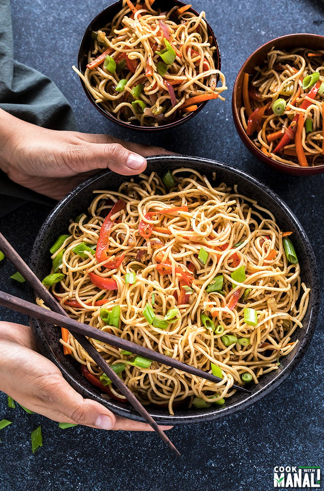 pair of hands holding a bowl of hakka noodles with chopsticks