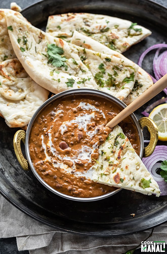 a piece of naan dipped in a kadai of dal makhani with more naan and onion slices in the plate