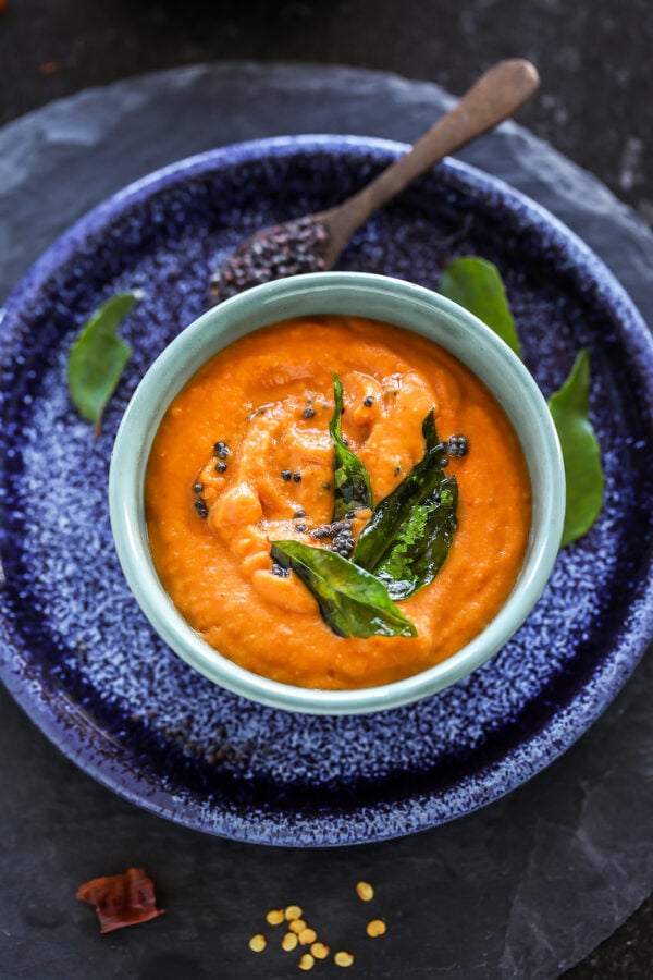 bowl of tomato chutney with curry leaves and mustard seeds thrown in the background
