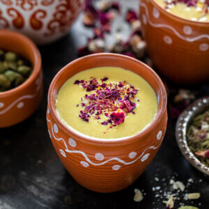 thandai in a small clay pot with small bowls with rose petals, cardamom on the sides and more clay pots of thandai in the backround