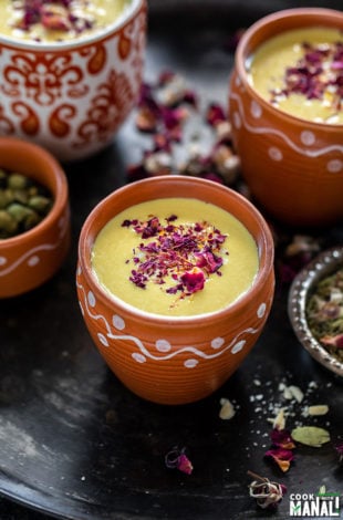 thandai in a small clay pot with small bowls with rose petals, cardamom on the sides and more clay pots of thandai in the backround