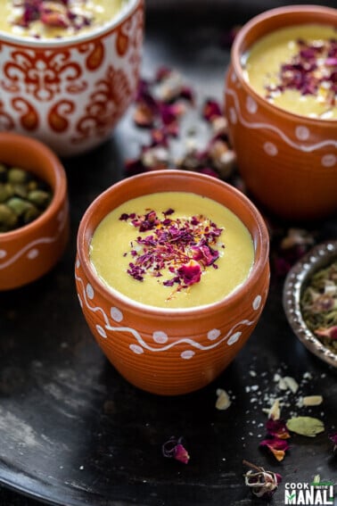 thandai in a small clay pot with small bowls with rose petals, cardamom on the sides and more clay pots of thandai in the backround