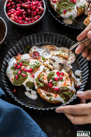 two aloo tikki served in a black rimmed plate and covered with yogurt, chutney and garnished with cilantro and pomegranate seeds