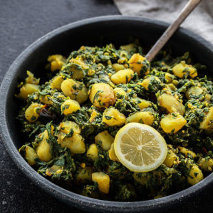 aloo palak in a black bowl with a spoon and served with a lemon wedge