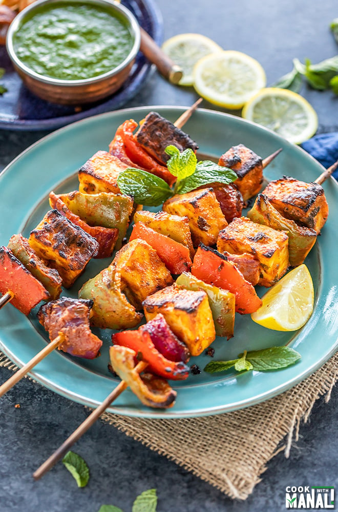 paneer tikka skewers arranged on a blue plate with lemon wedge on the side and bowl of cilantro chutney in the background