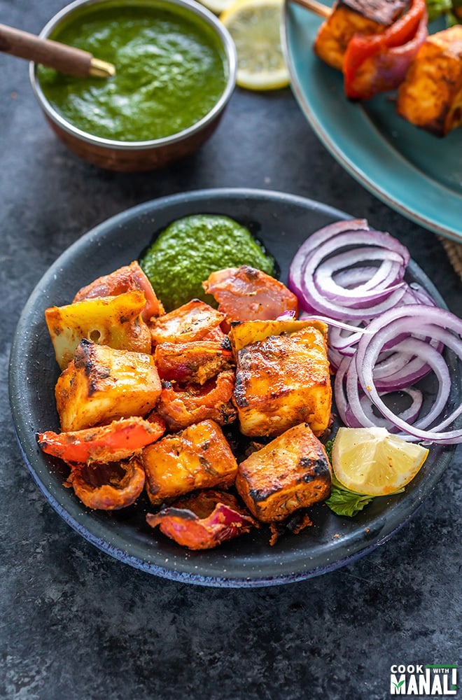 paneer tikka served with onion slices, cilantro chutney and slice of lemon in a black round plate