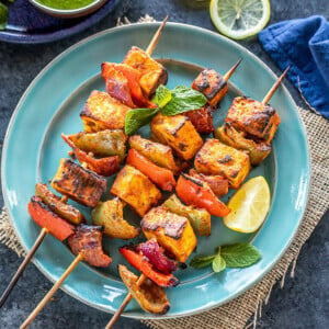 paneer tikka skewers arranged on a blue plate with lemon wedge on the side and bowl of cilantro chutney in the background