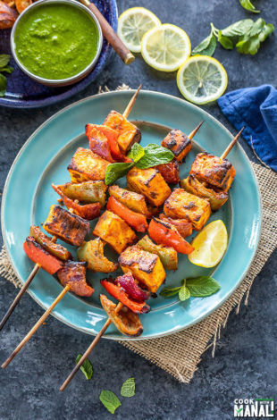 paneer tikka skewers arranged on a blue plate with lemon wedge on the side and bowl of cilantro chutney in the background