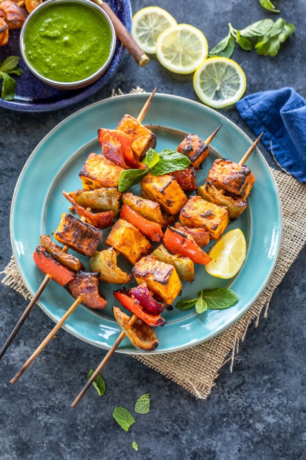 paneer tikka skewers arranged on a blue plate with lemon wedge on the side and bowl of cilantro chutney in the background