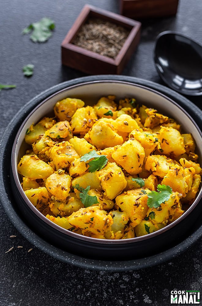 jeera aloo in a black bowl with a ladle and spice containers in the background