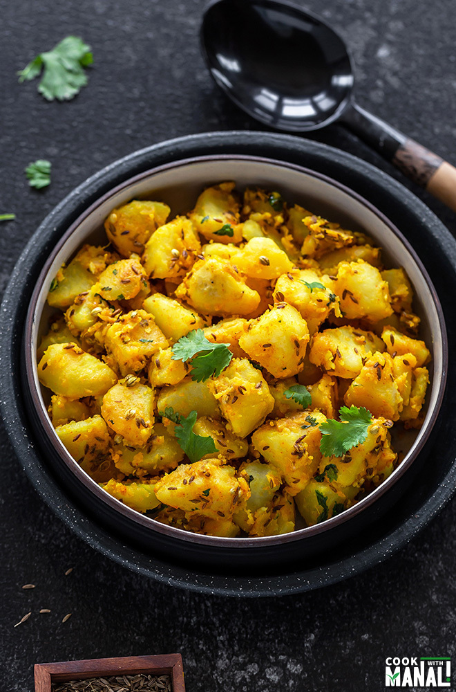 jeera aloo in a black bowl with a black wooden ladle on the side