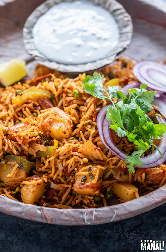 tawa pulao served in a plate garnished with onion rings, cilantro and served with a bowl of yogurt