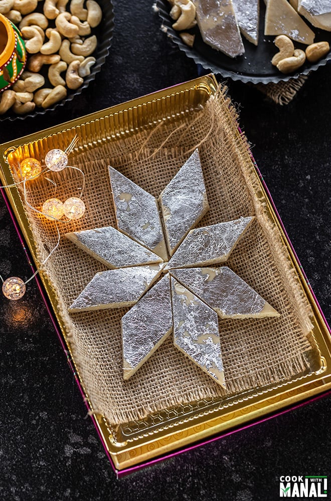 kaju katli arranged in a sweet box with small lights on the side and plate of cashews in the background