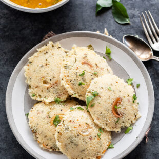 5 pieces of rava idli arranged in a plate with a bowl of sambar in the background
