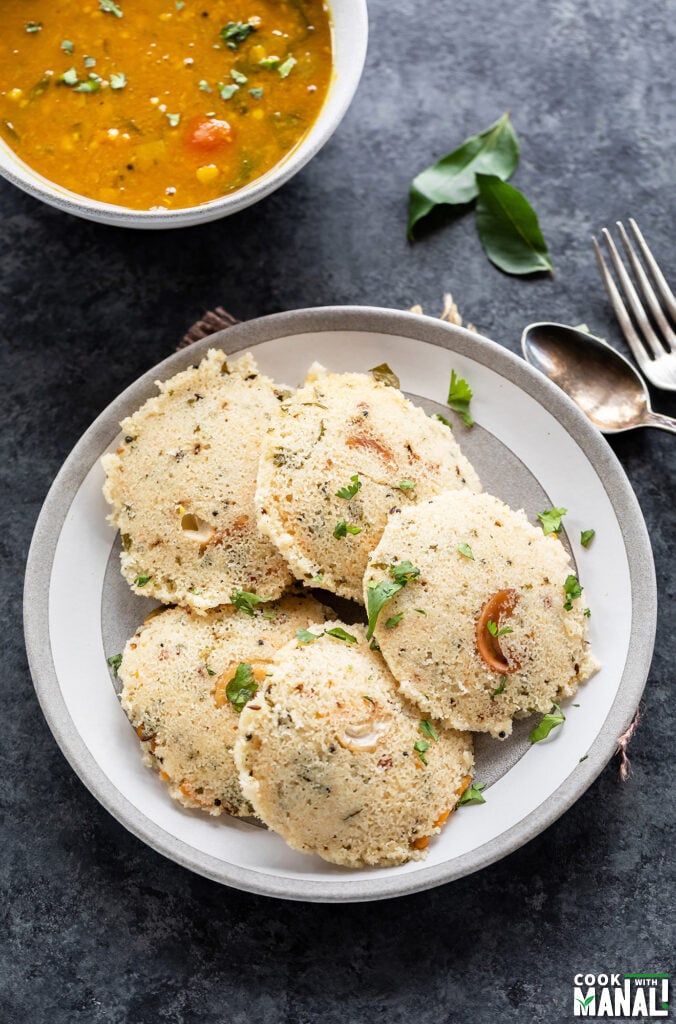 5 pieces of rava idli arranged in a plate with a bowl of sambar in the background