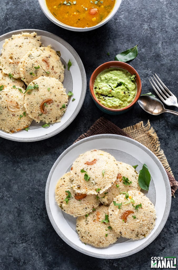 2 plates of rava idli with a bowl of coconut chutney and bowl of sambar in the background