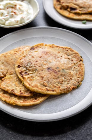 3 aloo parathas on a white plate with a bowl of yogurt in the back