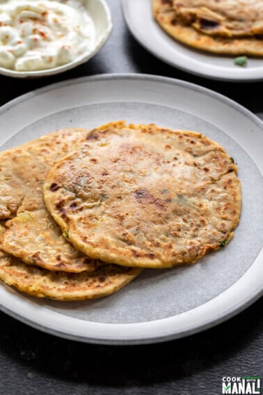 3 aloo parathas on a white plate with a bowl of yogurt in the back