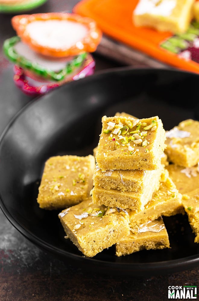 besan burfi stacked together on a black bowl with some diyas placed in the background