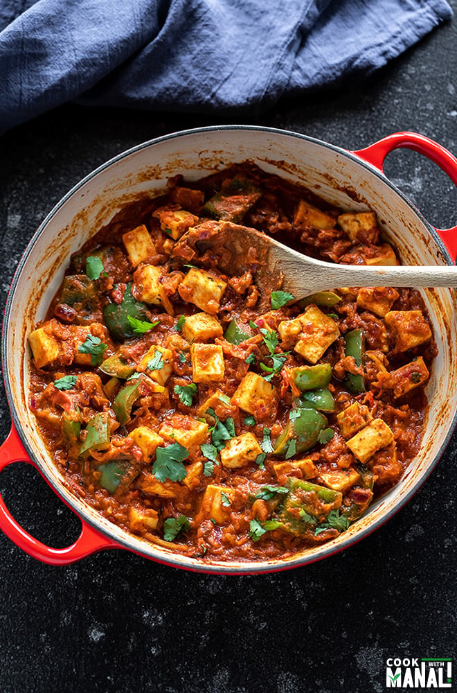 kadai paneer in a red color wok with a wooden spatula with blue napkin in the background
