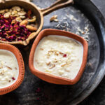 Rice Kheer served in a clay bowl and garnished with rose petals and a golden cast iron pan with nuts and rose petals in the background