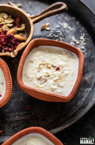 Rice Kheer served in a clay bowl and garnished with rose petals and a golden cast iron pan with nuts and rose petals in the background